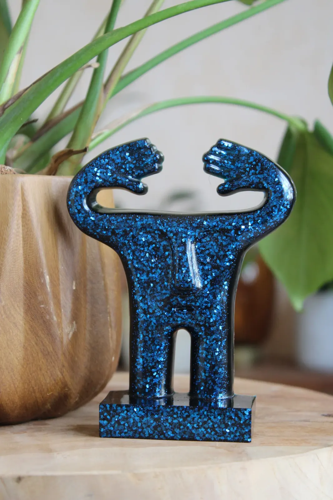 Statuette stylisée en résine verte pailletée aux bras levés posée près d'une plante