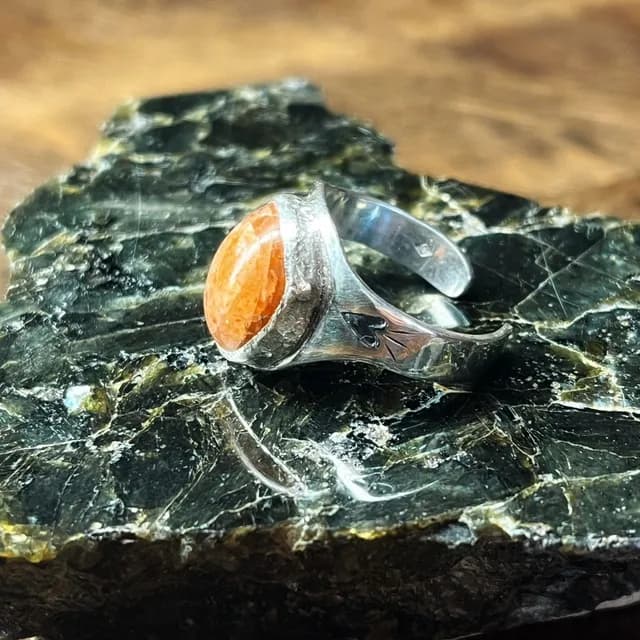 Bague chevalière en argent ornée d'une pierre de soleil orangée ovale posée sur une roche minérale.