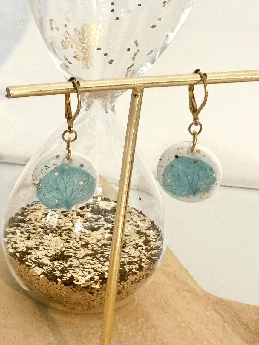 Boucles d'oreilles dormeuses dorées avec pampilles blanches et hortensia bleu présentées sur un porte-bijoux doré.