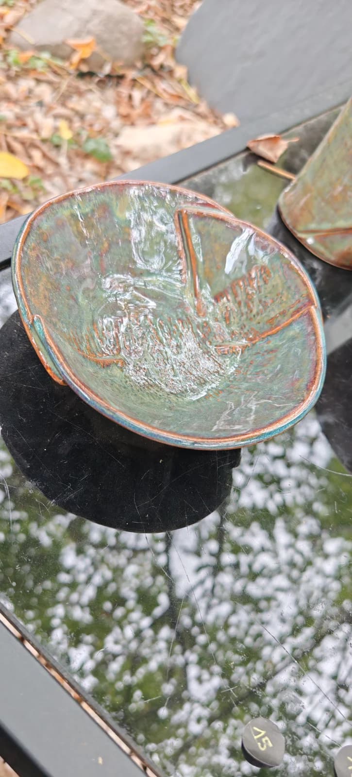 Coupelle en céramique émaillée verte et cuivrée avec empreinte végétale posée sur une table en verre.